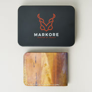 Hand-painted full-grain leather wallet with abstract art in purple, amber, and brown, displayed below a branded packaging tin. Markore Canvas Chaos.