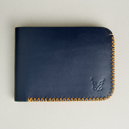 Front view of dark blue full-grain leather bifold wallet featuring handmade amber cross-stitched detailing on the edges and embossed logo. Markore Hunza Edge.