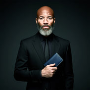 Studio portrait of a distinguished man with a grey beard and Vitiligo on his face wearing a black suit, holding the Markore Karakoram II in Indigo Blue close to his chest. The dark background and sharp lighting highlight both the man’s presence and the refined craftsmanship of the wallet.