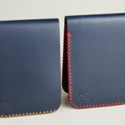 Side-by-side view of two dark blue full grain leather bifold wallets, featuring cross-stitched detailing in amber and red on the edges, and embossed logo. Markore Hunza Edge.