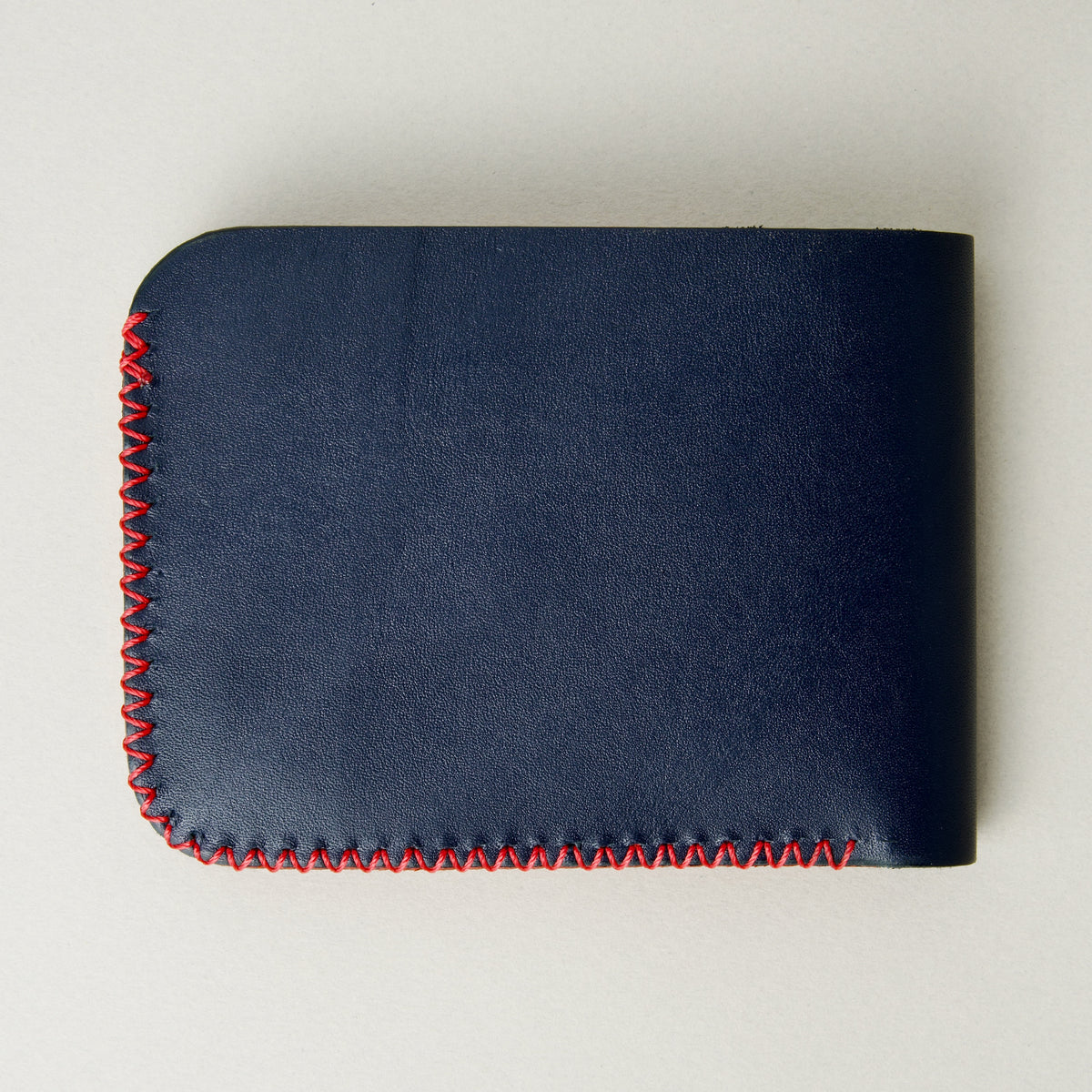 Back view of dark blue full-grain leather bifold wallet featuring red handmade cross-stitched detailing on the edges. Markore Hunza Edge.