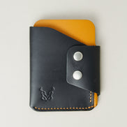 Front view of full grain leather card holder in black and amber, featuring side flap with two stainless steel press buttons, amber handmade stitching and embossed logo. Markore Niva Glide.
