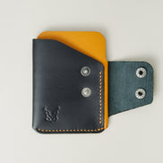 Front view of full grain leather card holder in black and amber, featuring an open side flap with two stainless steel press buttons, amber handmade stitching and embossed logo. Markore Niva Glide.