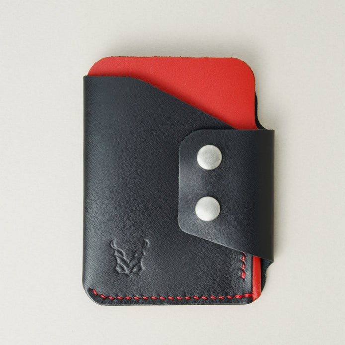 Front view of full grain leather card holder in black and red, featuring side flap with two stainless steel press buttons, red handmade stitching and embossed logo. Markore Niva Glide.