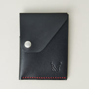 Front view of full grain leather card holder in black with red handmade stitching, featuring a flap with stainless steel press button and embossed logo. Markore Niva Heritage.