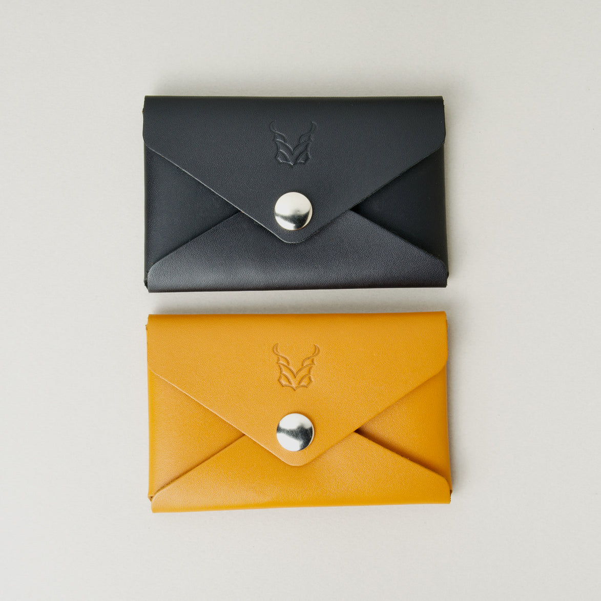 Side-by-side view of two full grain envelope style wallets in midnight black and amber with stainless steel press button and embossed logo. Markore Niva Seal
